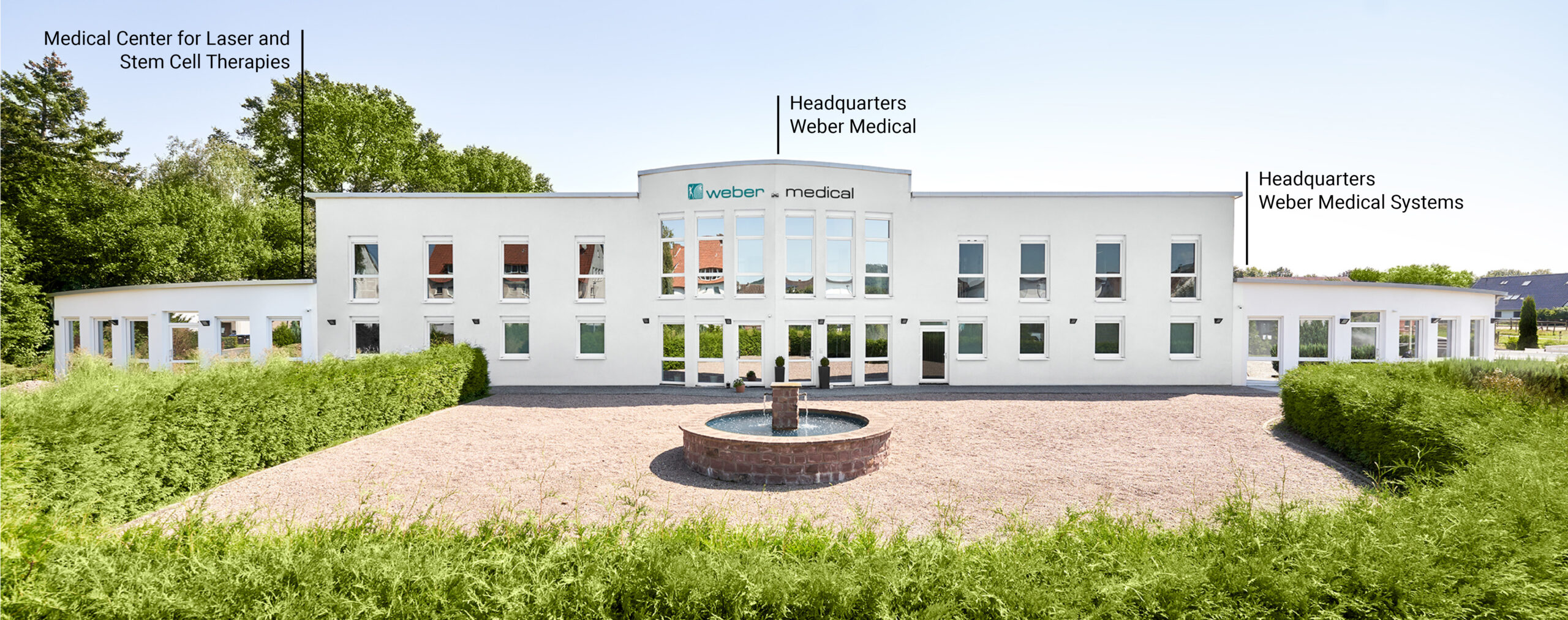 A white, two-story building with the sign Weber Medical above the entrance, set behind a circular driveway and a fountain, with trees and labeled company offices in the background.